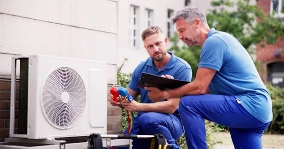 Two technicians checking an outdoor heat pump with pressure gauges and a tablet